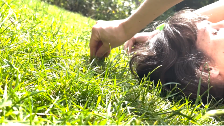 This close-up screen capture from a portal video shows bright sunlight on a field of green grass. Elisabeth Motley lays on her back, reaching her arms above her head to grasp blades of grass in her fingers, eyes closed in the sun. Photo: Dr. Petra Kuppers 