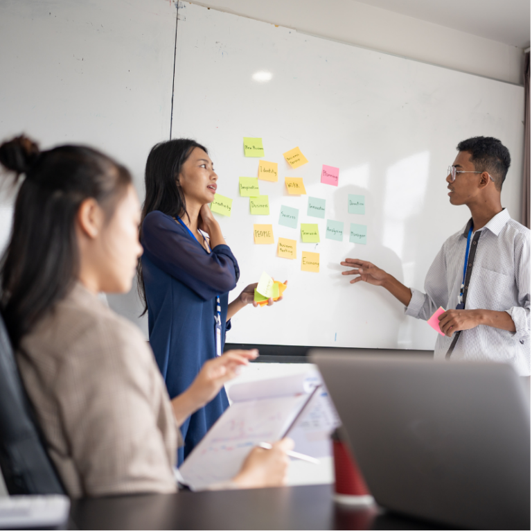 Three people collaborating using sticky notes, a notepad, and a computer.