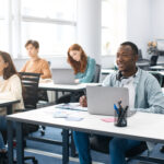 Education, Technology, Lifestyle And People Concept. Group of diverse international students sitting at desk in classroom using laptop computers, studying and listening to teacher, writing in notepads