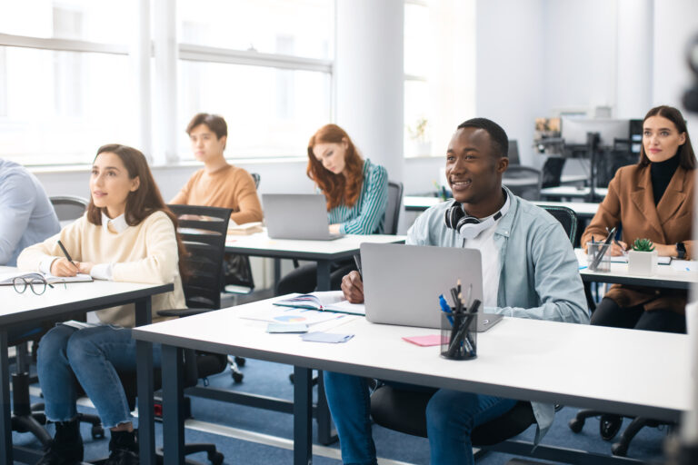 Education, Technology, Lifestyle And People Concept. Group of diverse international students sitting at desk in classroom using laptop computers, studying and listening to teacher, writing in notepads
